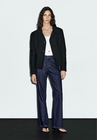 Svart ullblazer med knappar, vit linne, och mörkblå utsvängda jeans, kombinerat med platta sandaler. Modellen står mot en ljus bakgrund.