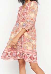 Blommönstrad klänning i nyanser av rosa och kräm, med långa ärmar och en flerlagers nederkant. Lättviktsmaterial med en mjuk textur.