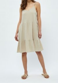 Ljus beige ärmlös klänning i ett texturerat tyg, med en utsvängd silhuett och en lager-på-lager fåll och fina band. Bärs med platta sandaler.