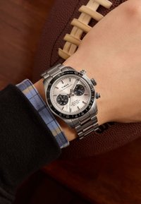 Sudraba nerūsējošā tērauda pulkstenis ar baltu ciparnīcu, melniem apakšciparnīciem, takymetrisko rāmīti un metāla siksniņu, kas uzlikts uz rokas, tuvumā pie futbola.