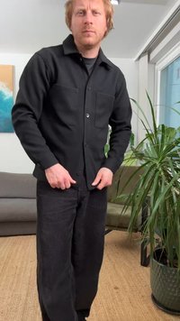 Camicia nera abbottonata con due tasche sul petto, abbinata a pantaloni larghi in velluto a coste neri. Il tessuto appare strutturato, con una vestibilità casual.