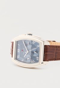 Ανδρικό ρολόι χειρός με ορθογώνιο ασημί καντράν, μπλε καντράν με ρωμαϊκούς αριθμούς, τρία υποκαντράν και καφέ δερμάτινο λουράκι.