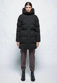 Schwarze Daunenjacke mit hohem Kragen und Kapuze, ausgestattet mit einem gesteppten Design, elastischem Bund und seitlichen Taschen. Mit grauen Hosen und Stiefeln kombiniert.