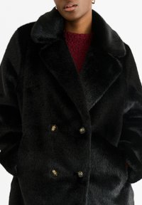 Double-breasted black faux fur coat with a wide collar and gold-tone buttons. The coat features a soft texture and a relaxed fit.