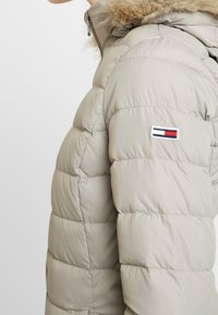 Lichtbeige pufferjack met horizontaal gestikt patroon, met een van bont voorziene capuchon en een klein logo-opdruk op de linkerschouder. Met een rits aan de voorkant.