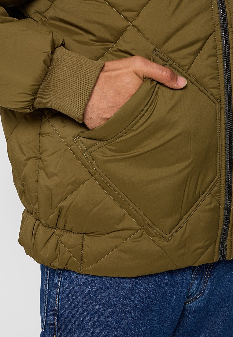 Olivgrüne gesteppte Jacke mit diagonaler Naht, gerippten Bündchen und Fronttasche. Verfügt über einen Reißverschluss und ein elegantes Design.
