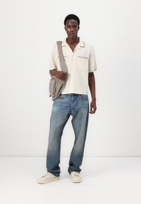 Short-sleeve cream shirt with black piping, light blue jeans, and beige sneakers, accompanied by a gray shoulder bag.
