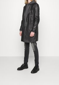Abrigo de cuero negro con un diseño elegante, mangas largas, cierre frontal y bolsillos; combinado con una sudadera gris y jeans ajustados. Zapatos negros.