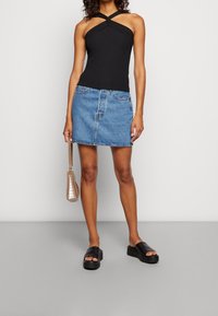 Mujer vestida con un top sin mangas negro de halter, una minifalda de mezclilla azul, sandalias de plataforma negras y sosteniendo un pequeño bolso beige.