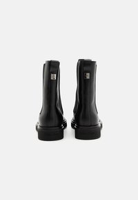 Botas de cuero negro hasta el tobillo con paneles laterales elásticos, acentos metálicos con logotipo y un tacón bajo en bloque, con una textura suave y brillante.
