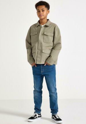 Poika seisoo kädet taskuissa, yllään beige takki, siniset farkut ja mustavalkoiset tennarit, yksivärisen valkoisen taustan edessä.