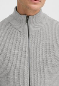 Lys grå zip-up genser med høy krage, med ribbestruktur og metallglidelås. Stoffet ser mykt og strikket ut.