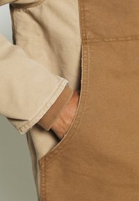 Hand in een bruine zak met een beige jasje zichtbaar, met een geribde manchet die de textuur van de stof en de stikdetails toont.