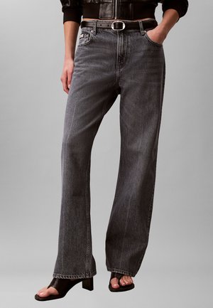 Personne portant un jean noir délavé taille haute à jambes larges, une ceinture en cuir noire, des chaussures noires à bout ouvert à talons blocs et une veste courte en cuir noir.