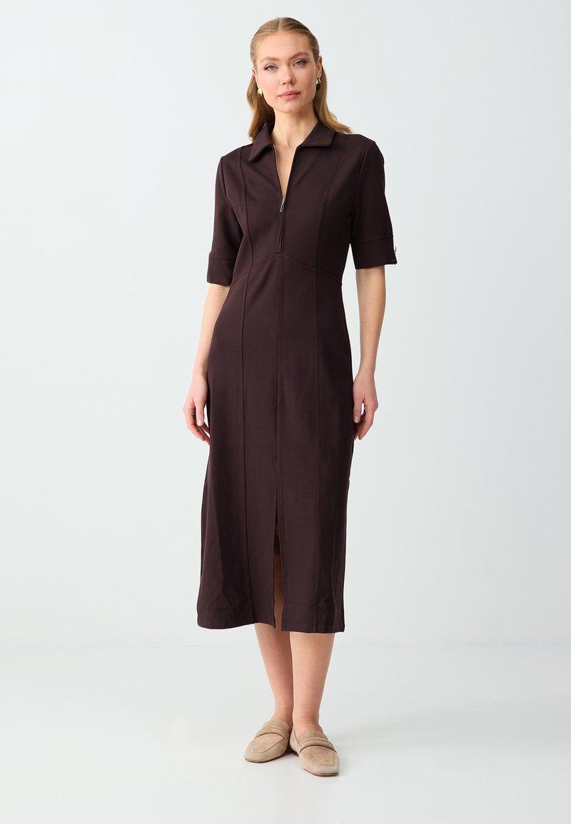 Femme debout dans une robe midi marron avec une fermeture éclair devant, des manches courtes, un col et des chaussures beige à enfiler sur un fond uni.