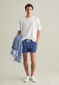 T-shirt bianco in cotone, pantaloni da bagno blu a righe, e un asciugamano a quadri blu. Sneakers bianche visibili, in piedi su uno sfondo neutro.