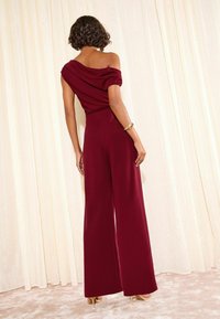 Salopeta burgundă are un design drapat cu umerii goi, o siluetă cu picioare largi și un material elastic, neted, fiind asortată cu o brățară aurie minimală.