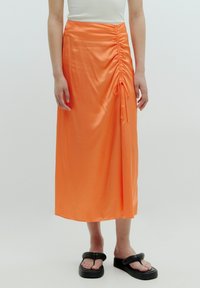 Mid-length oranjesatinrock mit gerafftem Detail und verstellbarem Bindebandverschluss an der Seite. Getragen mit schwarzen Plateausandalen.