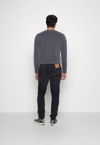 Grå långärmad skjorta, svarta jeans med bakfickor och blå sneakers med gula accenter. Modell sedd från baksidan, enkel bakgrund.