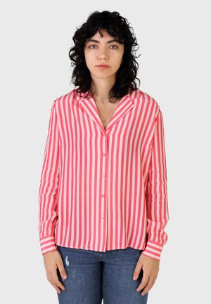 Kobieta o kręconych ciemnych włosach, ubrana w różowo-białą bluzkę z pionowymi paskami i niebieskie dżinsy, stojąca na jednolitym tle.