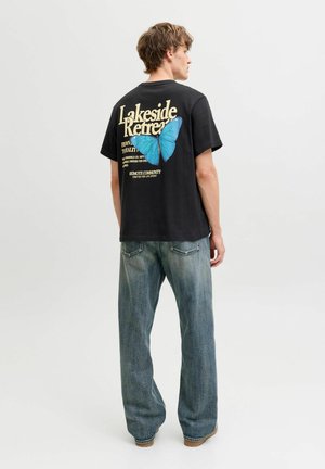 Homme vu de dos portant un t-shirt noir avec un grand papillon bleu et le texte « Lakeside Retreat », associé à un jean bleu ample.