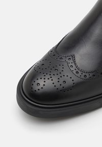 Chaussure en cuir noir à bout arrondi avec des motifs brogues perforés détaillés et une semelle en caoutchouc texturée. Finition lisse et accents de couture.