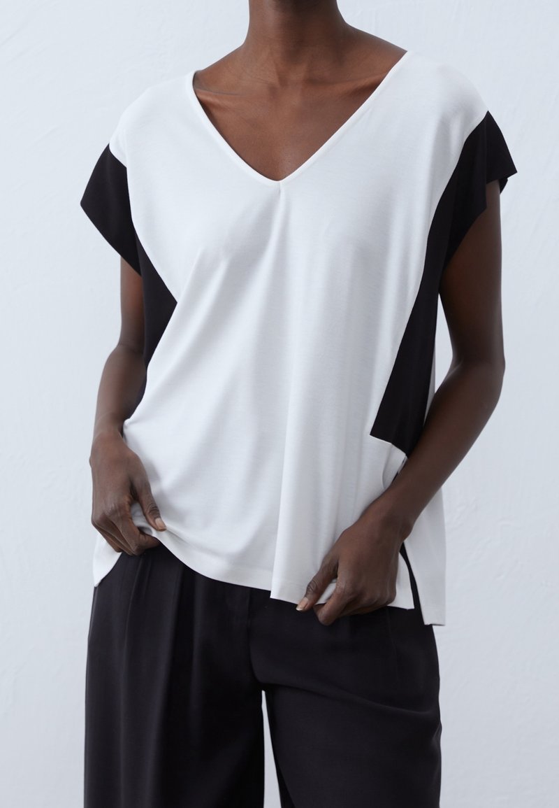 Top de manga corta con escote en V en diseño bicolor blanco y negro. Tela suave con un ajuste relajado. Aberturas laterales visibles en el dobladillo para mayor comodidad.