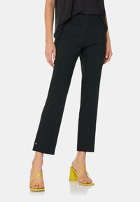 Zwarte broek met een rechte pasvorm en een patroon, voorzien van zij-splits en kleine zilveren hardware accenten, in combinatie met gele haksandalen.