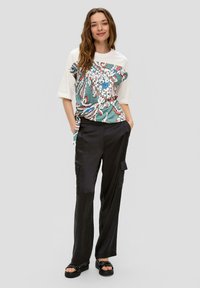 Hemelblauw, groen, rood en blauwe bloemetjespatroon op een shirt met witte mouwen. Zwarte cargobroek met zijzakken. Model draagt zwarte sandalen.