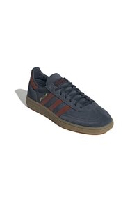 Sneaker in camoscio grigio con dettagli in pelle marrone, presenta tre strisce, lacci piatti e suola in gomma naturale texturizzata.