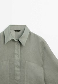 Light green linen shirt with a folded collar, buttoned placket, and two front pockets. Features a textured fabric and relaxed fit.