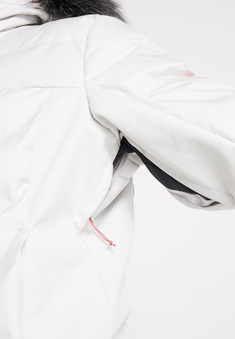Veste d'hiver blanche avec panneau de ventilation noir sous les aisselles et tirette de fermeture éclair rose, portée par une personne avec capuche bordée de fourrure visible.