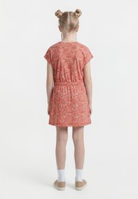 Ung pige med blondt hår sat op i rum-boller, iført en koralfarvet kortærmet blomsterkjole, hvide sokker og beige sko, stående med ryggen mod kameraet.