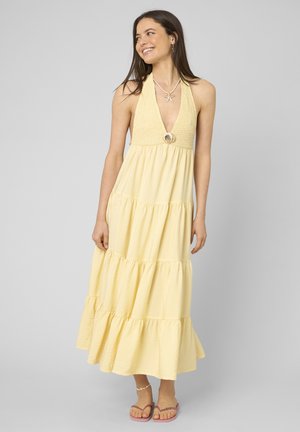 Femme souriante portant une robe longue jaune sans manches à étages, un collier de coquillages et des sandales roses, debout devant un fond gris uni.