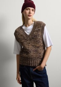 Maglione senza maniche marrone a maglia con scollatura a V, caratterizzato da un tessuto a trama sciolta e un motivo testurizzato, indossato sopra una T-shirt bianca e jeans scuri.