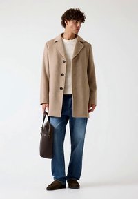 Beige wollen jas met een brede kraag en zwarte knopen, gecombineerd met een wit gestructureerde trui en een blauwe spijkerbroek. Bruine tas in de hand.