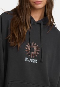 Czarna bluza z kapturem z kieszenią kangurką, z zakrzywioną grafiką pomarańczowego promienia słońca oraz napisem "BILLABONG" w białym kolorze z przodu.