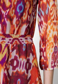 Cette robe à motifs présente des couleurs vives d'orange, de rouge, de violet et de bleu, avec une taille élastiquée et des manches bouffantes. Tissu lisse et léger.