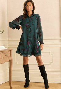 Rochie verde cu imprimeu floral, cu mâneci lungi transparente, guler negru și tiv în straturi, asortată cu cizme negre până la genunchi.