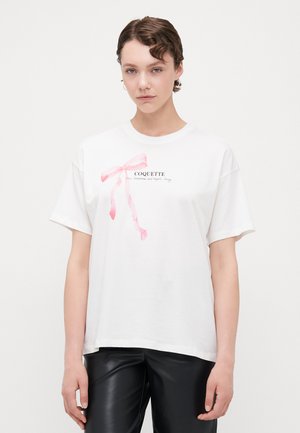 T-shirt en coton blanc à manches courtes, avec un graphique de ruban rose et le mot "COQUETTE" en texte noir en dessous.