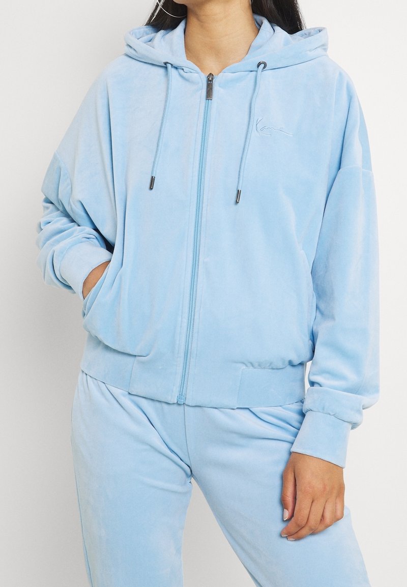 Femme portant un sweat à capuche bleu clair zippé et un pantalon assorti, une main dans la poche, sur fond blanc uni.