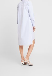 Žena ve modro-bílém pruhovaném dlouhém triku a bílých sandálech na vysokém podpatku, stojící na jednolitém pozadí.