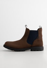 Stivaletto in suede marrone con pannelli laterali elasticizzati blu navy, linguette e suola in gomma testurizzata. Presenta una punta arrotondata e cuciture minime.