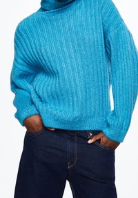 Turquoise ribbed sweater with a high neck, featuring a soft texture and relaxed fit, paired with dark blue denim jeans.