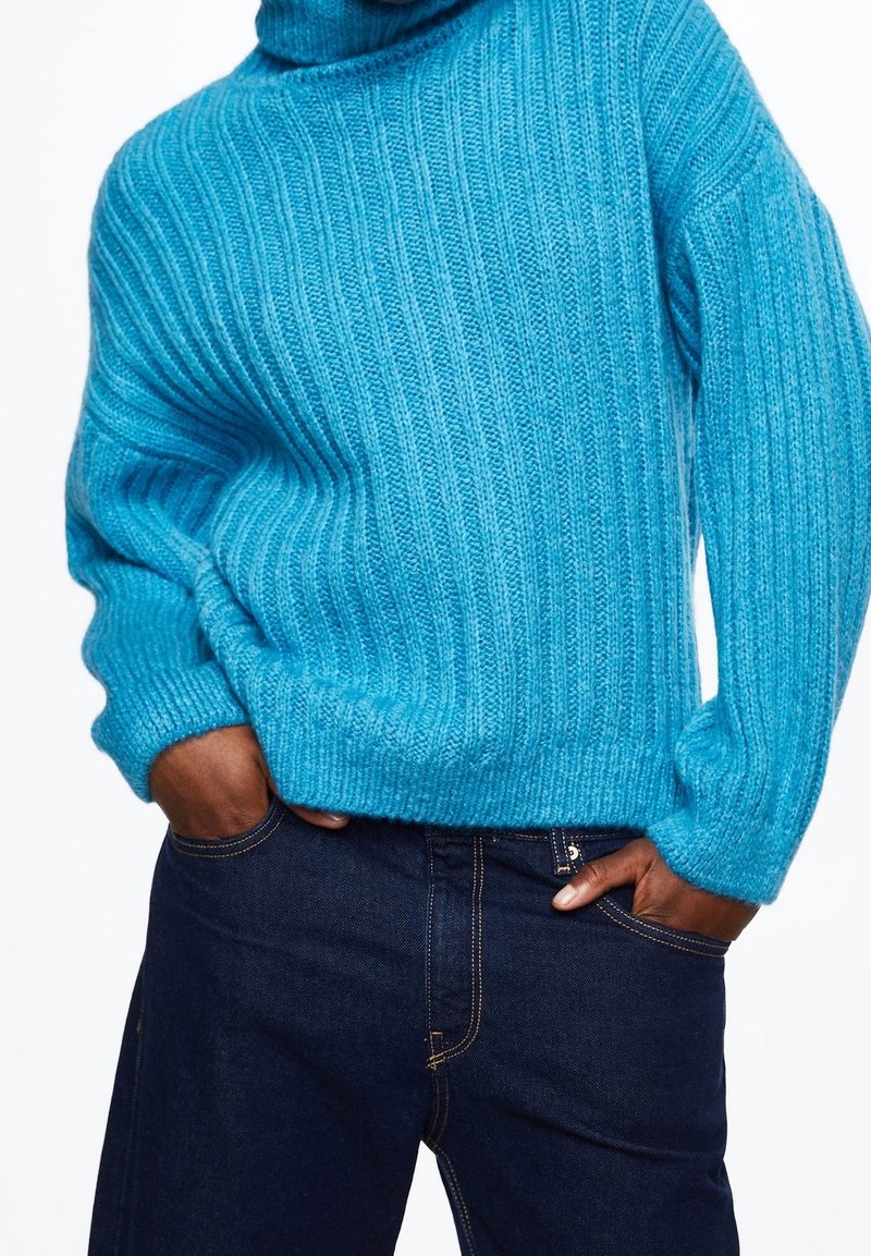 Turquoise ribbed sweater with a high neck, featuring a soft texture and relaxed fit, paired with dark blue denim jeans.