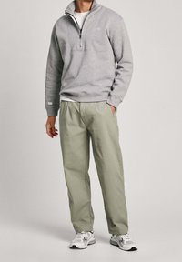 Felpa grigia con zip a quarto e tasca frontale, abbinata a pantaloni verde chiaro. Il modello indossa scarpe da ginnastica bianche e una maglietta bianca sotto.