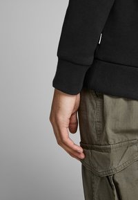 Black sweatshirt with ribbed cuffs, paired with olive green cargo pants featuring side pockets and textured fabric. Hand resting on the leg.