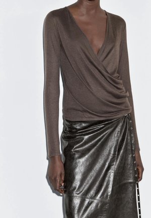 Femme portant un haut cache-cœur marron à manches longues et une jupe en cuir noire taille haute avec des boutons sur le côté, sur fond blanc.