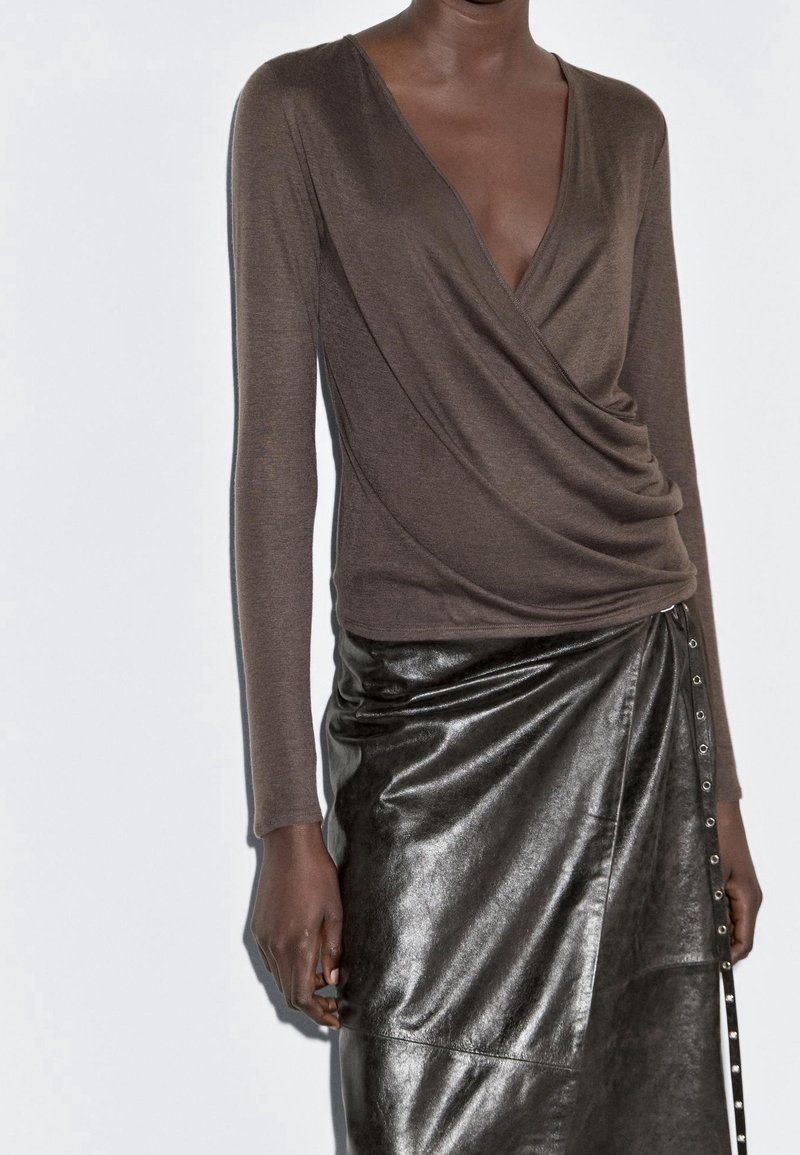 Femme portant un haut cache-cœur marron à manches longues et une jupe en cuir noire taille haute avec des boutons sur le côté, sur fond blanc.