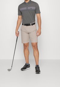 Muž v šedém golfovém polo, béžových šortkách, s černým opaskem a černými teniskami, drží golfovou hůl před čistým bílým pozadím.